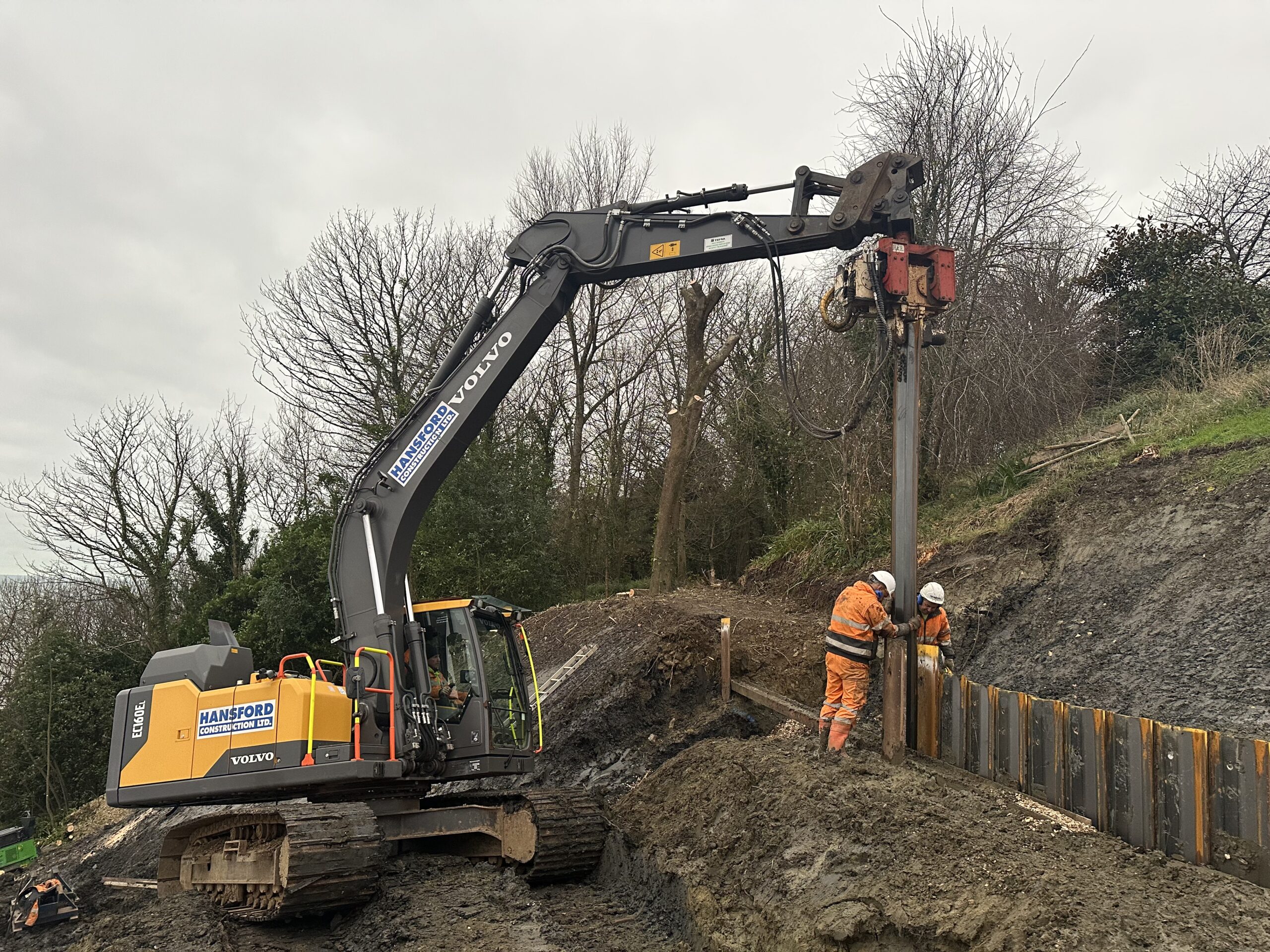 Excavator driving sheet piles to stabilise hillside and prevent further landslip in Lyme Regis